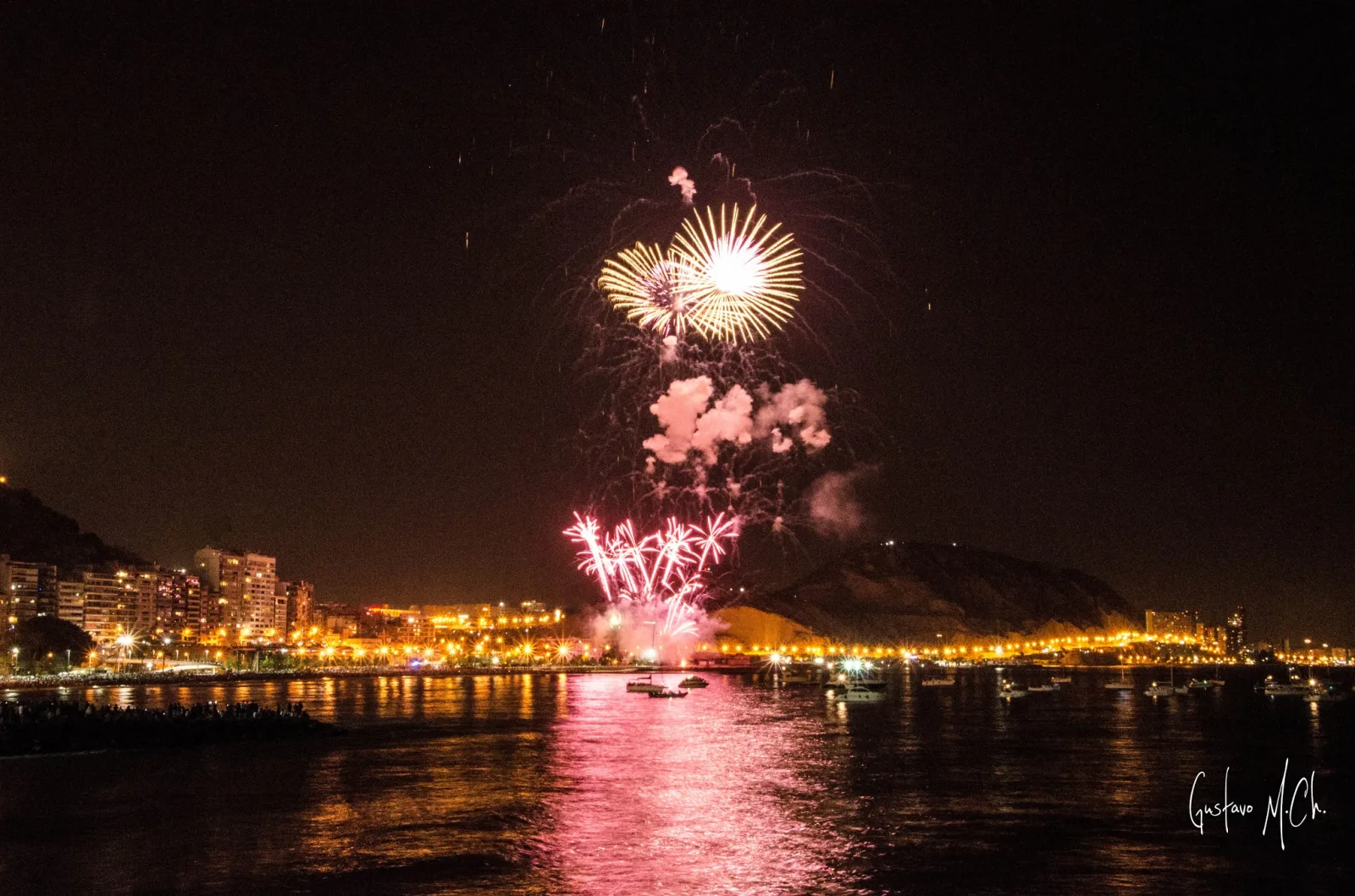 Fuegos artificiales sobre la costa iluminada de Alicante y reflejos en el mar